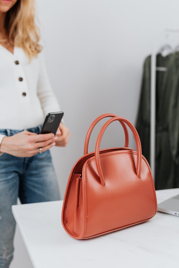 A woman in casual attire uses her phone to photograph an elegant leather handbag indoors.